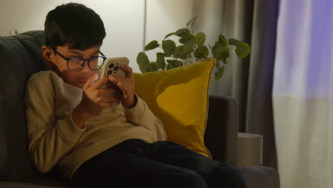 Young-Boy-Sitting-On-Sofa-In-Lounge-At-Home-Playing-Game-On-Mobile-Phone-At-Night
